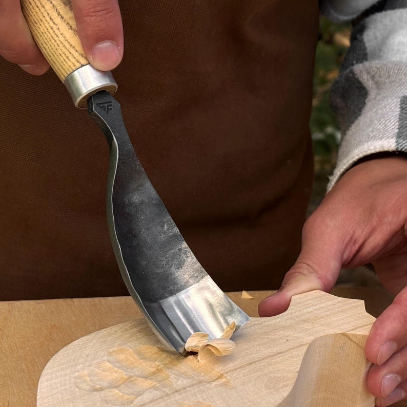 Pu&ograve; includere: Una sgorbia curva per intaglio del legno con una lama in metallo lucido e un manico in legno chiaro. L'utensile presenta un marchio distintivo del produttore vicino alla ghiera e rimuove un sottile truciolo di legno chiaro da una superficie.