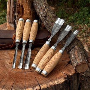 May include: A set of six wood carving gouges featuring light-colored wooden handles with visible grain patterns, metal ferrules, and curved steel blades. The tools are arranged in a group, with three smaller gouges and three larger gouges.