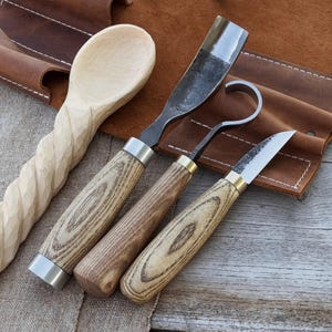 May include: A wooden spoon with a carved handle and three wood carving tools. The tools have wooden handles with metal blades and collars. The tools are a gouge, a hook knife, and a detail knife. The tools are displayed on a brown leather surface.