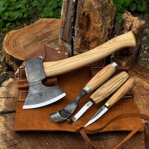 May include: A wood carving set featuring a hatchet with a dark metal head and light wood handle, a curved hook knife, a gouge, and a detail knife, all with matching light wood handles, arranged on a brown leather tool roll with a leather sheath.