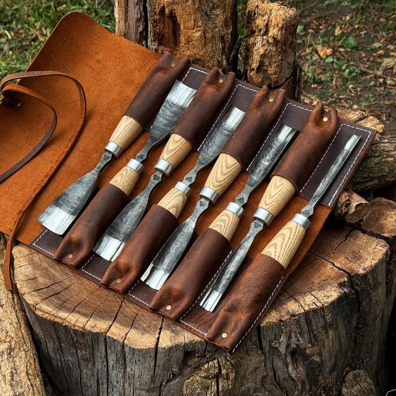 May include: A set of six wood carving chisels with light-coloured wooden handles and dark metal blades, organised in a brown leather roll-up tool pouch with white stitching. The tools feature varied blade shapes for detailed woodworking tasks.