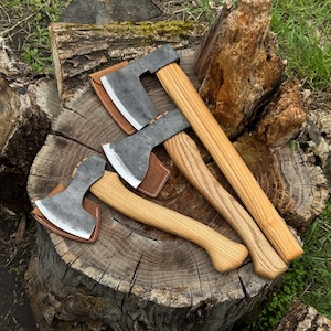 Peut inclure: Quatre haches avec des manches en bois et des lames en métal gris sont présentées sur une souche d'arbre. Chaque hache a un étui en cuir marron. Les manches en bois varient en longueur et en veinage.