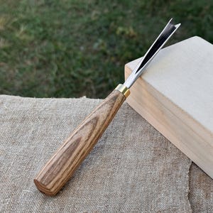 May include: A woodworking gouge with a wooden handle and a metal blade. The handle is a light brown color with a wood grain pattern. The blade is silver and black. The gouge is resting on a tan surface.