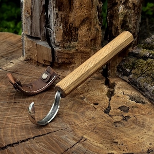 May include: A hook knife with a long, cylindrical wooden handle and a curved, polished metal blade. Beside the knife lies a small, brown leather sheath featuring a metal snap button closure and a stamped logo on the exterior surface.