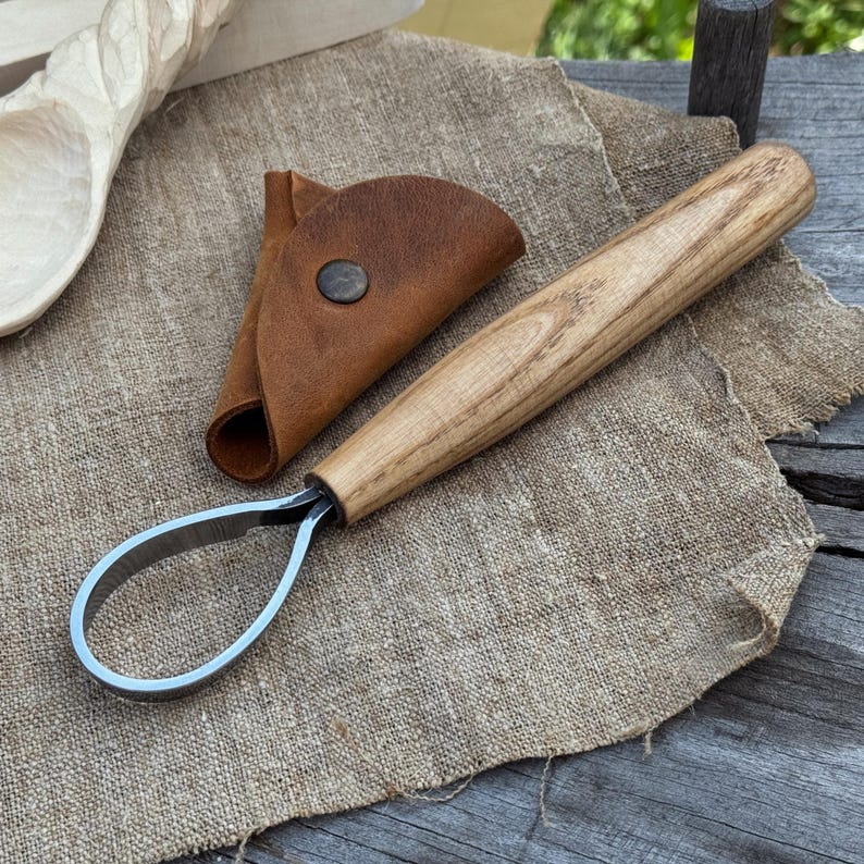 May include: A wood carving hook knife with a curved blade and a wooden handle, next to a brown leather sheath. The knife is designed for detailed wood carving and sits on a textured, neutral-colored fabric.