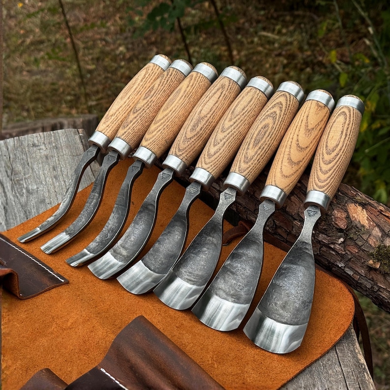May include: A set of eight wood carving gouges with light brown wooden handles and silver metal ferrules. Each tool features a curved, dark, forged steel blade with a polished edge, arranged in a row on a brown leather tool roll.