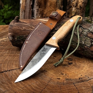 May include: A fixed-blade knife with a light-coloured wooden handle, a silver metal blade with a dark textured finish, and a green paracord lanyard. The knife includes a brown leather sheath with white stitching and a stamped logo on the surface.