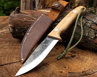 May include: A fixed-blade knife with a light-colored wooden handle, a silver metal blade with a dark textured finish, and a green paracord lanyard. The knife includes a brown leather sheath with white stitching and a stamped logo on the surface.
