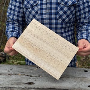 May include: A rectangular wooden panel with a geometric pattern of triangles and stars. The panel is light brown and held by a person wearing a blue plaid shirt. The design is etched into the wood.