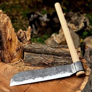 May include: A hand-forged, rustic-style draw knife with a wooden handle. The blade is dark gray with a sharp edge, and the handle is light-colored wood. The knife rests on a wooden surface, suggesting its use in woodworking or crafting.