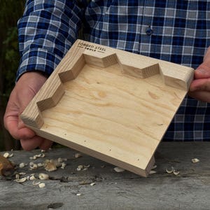 May include: A light-coloured wooden carving tray with a star-shaped edge. The tray is held by a person wearing a blue and black plaid shirt. The text "FORGED STEEL TOOLS" is visible on the tray.