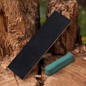 Op de afbeelding: Een zwarte slijpriem met een groene honen compound. De slijpriem is rechthoekig, gemaakt van zwart vilt en gemonteerd op een houten basis. De groene honen compound staaf is rechthoekig en ligt naast de slijpriem.