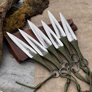 Peut inclure: Six couteaux de lancer à lames argentées, avec des manches enveloppés de vert olive et des longes attachées. Les lames sont tranchantes et pointues, et les manches ont des anneaux métalliques. Les couteaux sont disposés sur une surface beige avec une pochette en cuir.