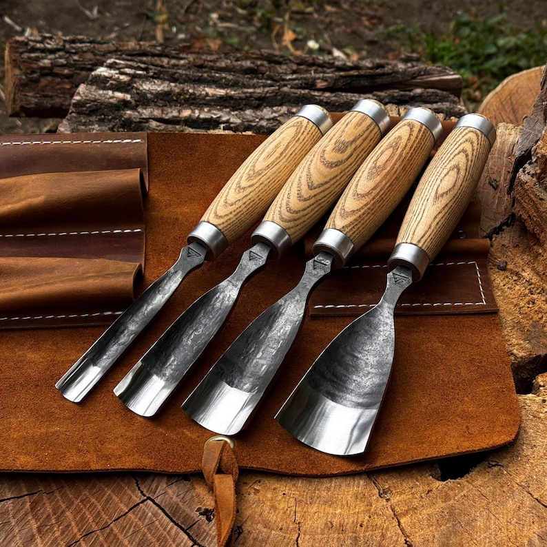 Puede incluir: Un juego de cuatro formones para madera con mangos de madera clara y virolas de metal plateado. Las hojas son de acero oscuro y texturizado con anchos y bordes curvos variables, dispuestos sobre un estuche de herramientas de cuero marr&oacute;n.