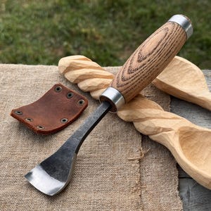 Puede incluir: Una herramienta para tallar madera con una hoja de metal oscura y curva, mango de madera clara y una virola de metal plateado. Junto a la herramienta hay una peque&ntilde;a funda de cuero marr&oacute;n con cuatro remaches met&aacute;licos y dos cucharas de madera sin terminar.