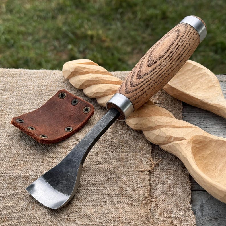 Puede incluir: Una herramienta para tallar madera con una hoja de metal oscura y curva, mango de madera clara y una virola de metal plateado. Junto a la herramienta hay una peque&ntilde;a funda de cuero marr&oacute;n con cuatro remaches met&aacute;licos y dos cucharas de madera sin terminar.