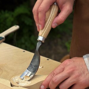 May include: A hook knife with a curved metal blade and a light-colored wooden handle. The tool is being used to carve a shallow depression into a piece of light-colored wood, creating small wood shavings as the blade removes material.