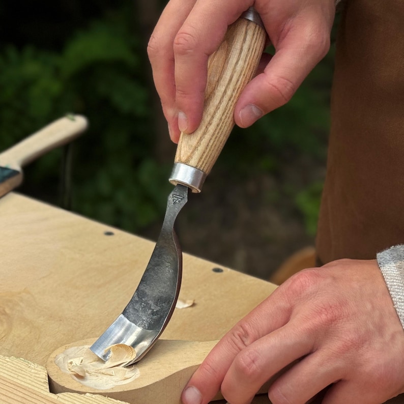 May include: A hook knife with a curved metal blade and a light-colored wooden handle. The tool is being used to carve a shallow depression into a piece of light-colored wood, creating small wood shavings as the blade removes material.
