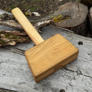 Puede incluir: Un mazo de madera con un mango marrón claro y una cabeza rectangular. La cabeza está hecha de varias piezas de madera unidas. El mazo está diseñado para carpintería o tareas similares.