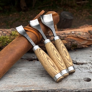 May include: Three wood carving tools with curved metal blades and light-colored wooden handles featuring a distinct grain pattern. Each tool has a silver metal ferrule connecting the blade to the handle, and they are arranged together.