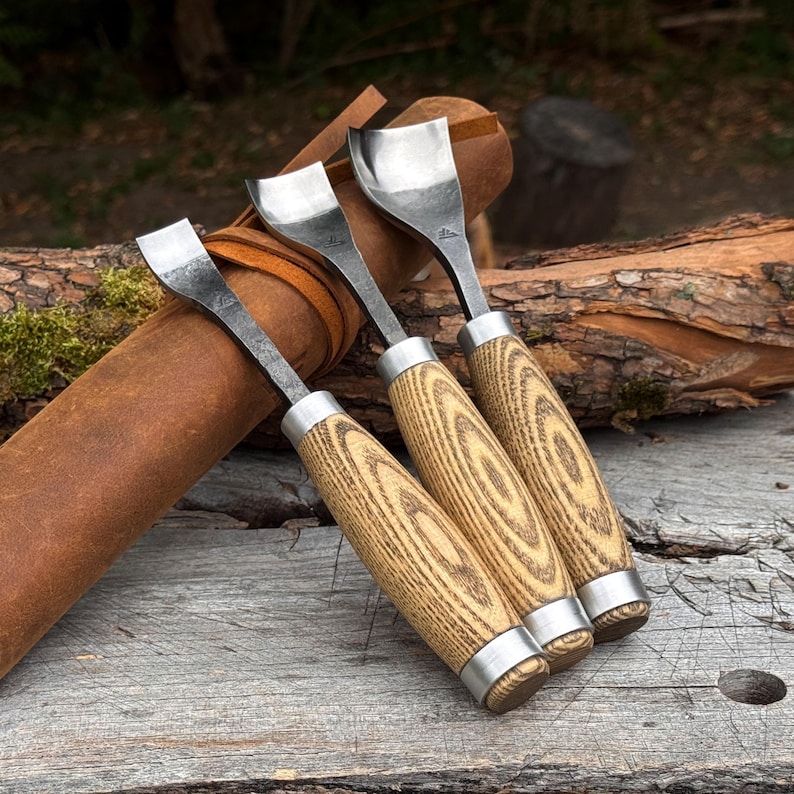 May include: Three wood carving tools with curved metal blades and light-colored wooden handles featuring a distinct grain pattern. Each tool has a silver metal ferrule connecting the blade to the handle, and they are arranged together.