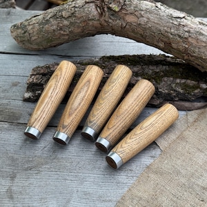 May include: Five wooden handles with a light brown finish and silver-coloured metal collars. The handles are arranged on a weathered wooden surface, with a piece of bark and burlap in the background. The handles have a cylindrical shape.