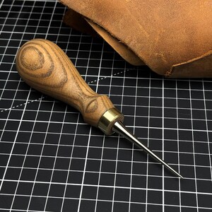 May include: A leatherworking awl with a wooden handle and a metal tip. The handle is light brown with a wood grain pattern. The awl is lying on a black and white grid surface, with a piece of brown leather in the background.