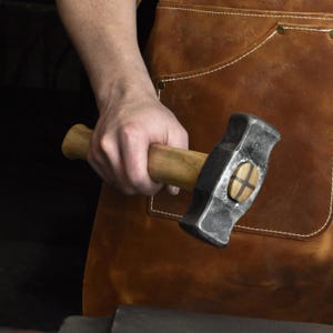 May include: A blacksmith's hammer with a wooden handle and a dark, textured head. The hammer's head features a decorative wooden inlay. The background includes a brown leather apron.
