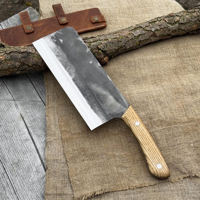 Puede incluir: Una gran cuchilla rectangular con un mango de madera clara y una hoja oscura y texturizada. El cuchillo descansa sobre una tela marr&oacute;n con una funda de cuero marr&oacute;n y un trozo de madera en el fondo. La hoja mide unos 25 cm de largo.