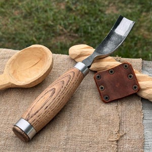 May include: A wood carving spoon gouge with a curved blade and a wooden handle. The handle is light brown with a silver ferrule. A brown leather sheath with metal rivets is next to the gouge. A carved wooden spoon is in the background.