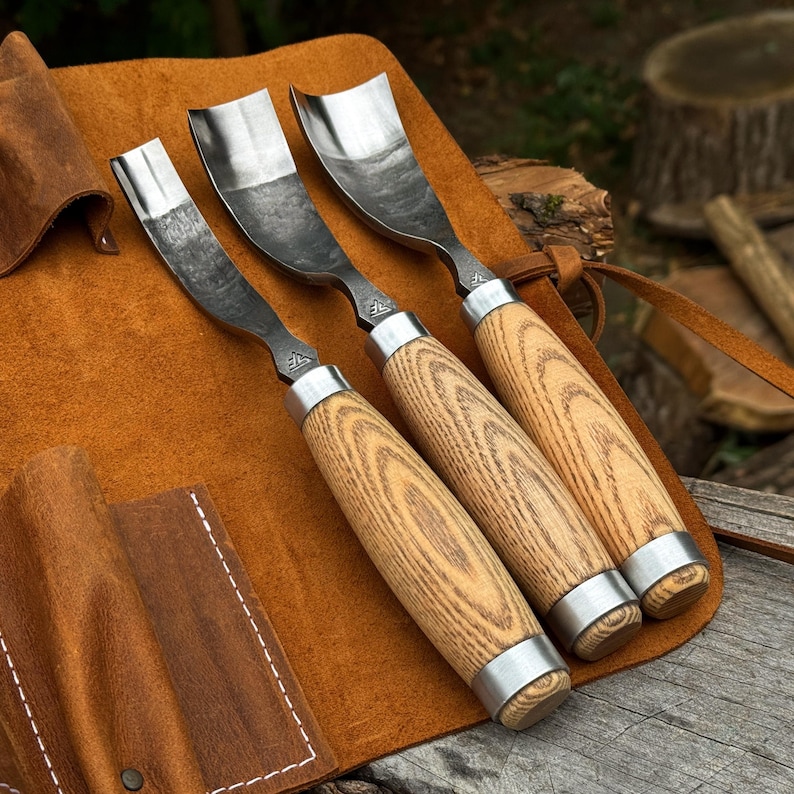 Puede incluir: Tres gubias para tallar madera con hojas de metal curvadas y mangos de madera clara. Cada herramienta presenta una virola de metal en la base del mango y un peque&ntilde;o logotipo estampado en el v&aacute;stago met&aacute;lico. Herramientas sobre un rollo de cuero.