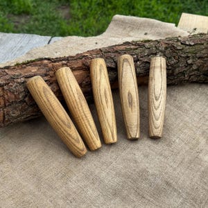 May include: Five wooden handles with a natural wood grain pattern. The handles are cylindrical with rounded ends and are arranged on a neutral-colored linen surface. A piece of bark rests behind the handles.