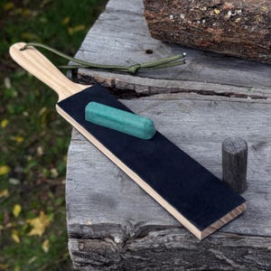 May include: A wooden strop with a black leather surface and a light green sharpening stone. The strop has a wooden handle and a green cord loop. A small wooden cylinder is also present.
