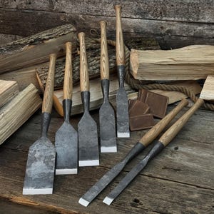 May include: A collection of seven hand tools featuring dark, forged metal blades with sharp, flat edges and long, light-colored wooden handles with visible grain patterns. The tools are arranged in a group, with some standing upright and others lying flat.