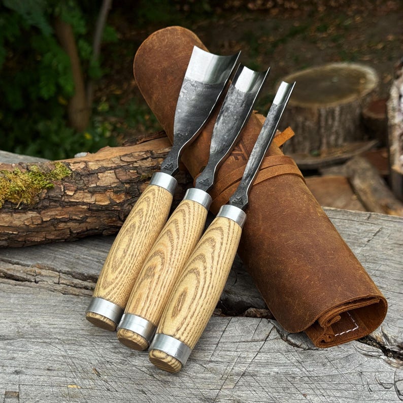 May include: Three wood carving gouges with curved steel blades and light-coloured wood handles featuring prominent grain patterns. Each tool has a silver metal ferrule at the base of the handle. The gouges are arranged together on a brown leather roll.