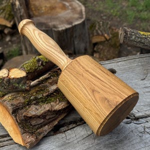 May include: A wooden mallet with a rounded handle and a rectangular head. The mallet is made of light-colored wood and rests on a weathered wooden surface with moss and tree bark in the background.