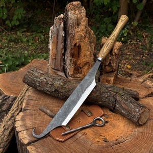 Könnte beinhalten: Ein handgeschmiedetes Messer mit einer dunklen, texturierten Stahlklinge, die an der Spitze einen gebogenen Haken aufweist, und einem hellen Holzgriff. Eine hellbraune Lederscheide mit Druckknopf und eine kleine Ringschraube liegen unter der Klinge.