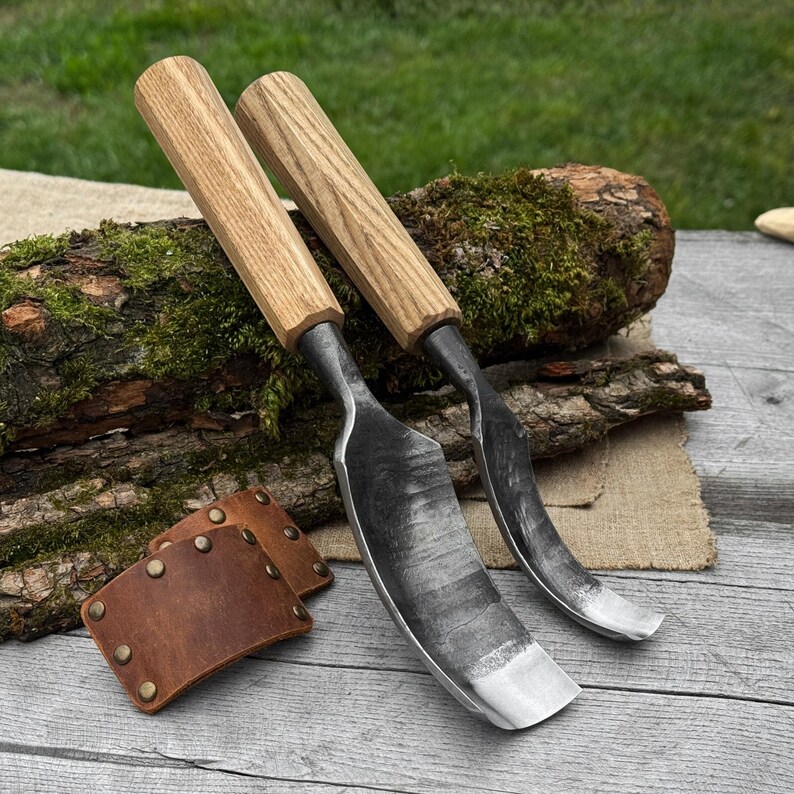 May include: Two wood carving gouges with wooden handles and curved steel blades. The blades are silver and the handles are light brown. A brown leather sheath with brass rivets is also present. The tools are designed for woodworking.