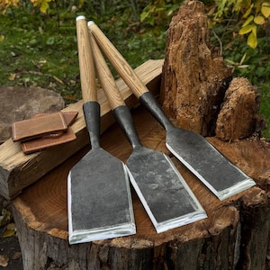 Può includere: Tre utensili manuali con lunghi manici in legno chiaro e lame larghe, piatte e in metallo grigio scuro. Gli utensili sono disposti fianco a fianco, caratterizzati da una finitura in metallo forgiato testurizzato e venature del legno visibili.