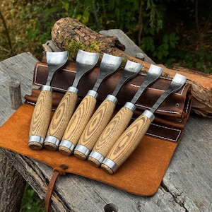 May include: A set of six wood carving gouges with curved metal blades and light-colored wooden handles with visible grain patterns. The tools are arranged in a row, resting on a brown leather tool roll with white stitching and metal rivets.