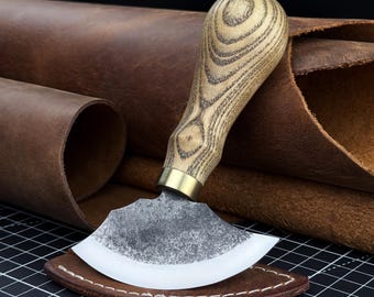 May include: A leathercraft head knife featuring a curved, sharpened steel blade with a textured finish, a brass-colored metal ferrule, and a light-colored wooden handle with visible grain patterns, resting on a brown leather piece.