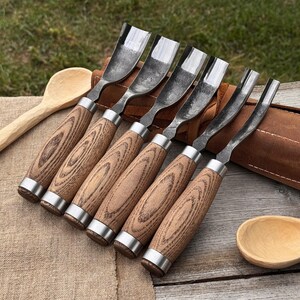 May include: A collection of six wood carving gouges, each with a wooden handle and a metal blade. The handles display a light brown wood grain and are finished with silver metal rings. A leather tool roll and a wooden spoon are also present.