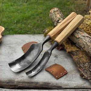 May include: Two wood carving gouges with curved, dark metal blades and light wood handles. The tools rest on a weathered wooden surface, accompanied by two small, brown leather blade protectors. The background features green grass and a piece of wood.
