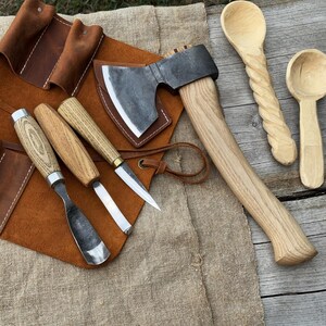 Puede incluir: Una colección de herramientas para trabajar la madera, que incluye un hacha, cuchillos para tallar y cinceles, exhibidos sobre un rollo de herramientas de cuero marrón y una superficie de madera. También hay dos cucharas de madera. Las herramientas tienen mangos de madera y hojas de metal.