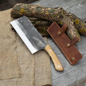 Puede incluir: Una gran cuchilla forjada a mano con un mango de madera clara y una funda de cuero. La hoja tiene un acabado oscuro y texturizado con un filo afilado. La funda de cuero marr&oacute;n tiene dos correas con broches.
