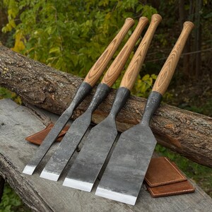 Puede incluir: Cuatro herramientas manuales con mangos largos de madera y hojas anchas, planas y oscuras de metal con bordes afilados. Las herramientas están dispuestas en fila, mostrando anchos de hoja variables, con fundas de cuero marrón cerca de la base.