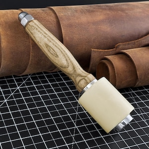 May include: A leathercraft mallet with a light wood handle and a cream-colored striking head. The mallet is set against a backdrop of rolled and flat brown leather hides, suggesting a leatherworking theme.
