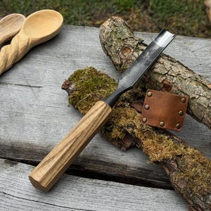 Op de afbeelding: Een houtsnijbeitel met een houten handvat en een gebogen metalen blad. De handgreep is lichtbruin met verticale groeven. Een kleine, bruine leren schede met messing klinknagels ligt naast de beitel. Twee houten lepels staan op de achtergrond.