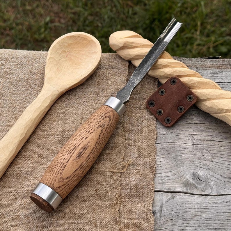 May include: A wood carving gouge with a polished metal blade and a light brown wooden handle with silver ferrules, positioned next to a hand-carved wooden spoon, a twisted wooden rod, and a small square brown leather sheath with metal rivets.