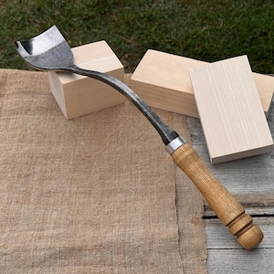 May include: A curved, metal carving tool with a wooden handle. The blade is silver and curved, and the handle is light brown. The tool is designed for woodworking and is shown with several small, unfinished wooden blocks.